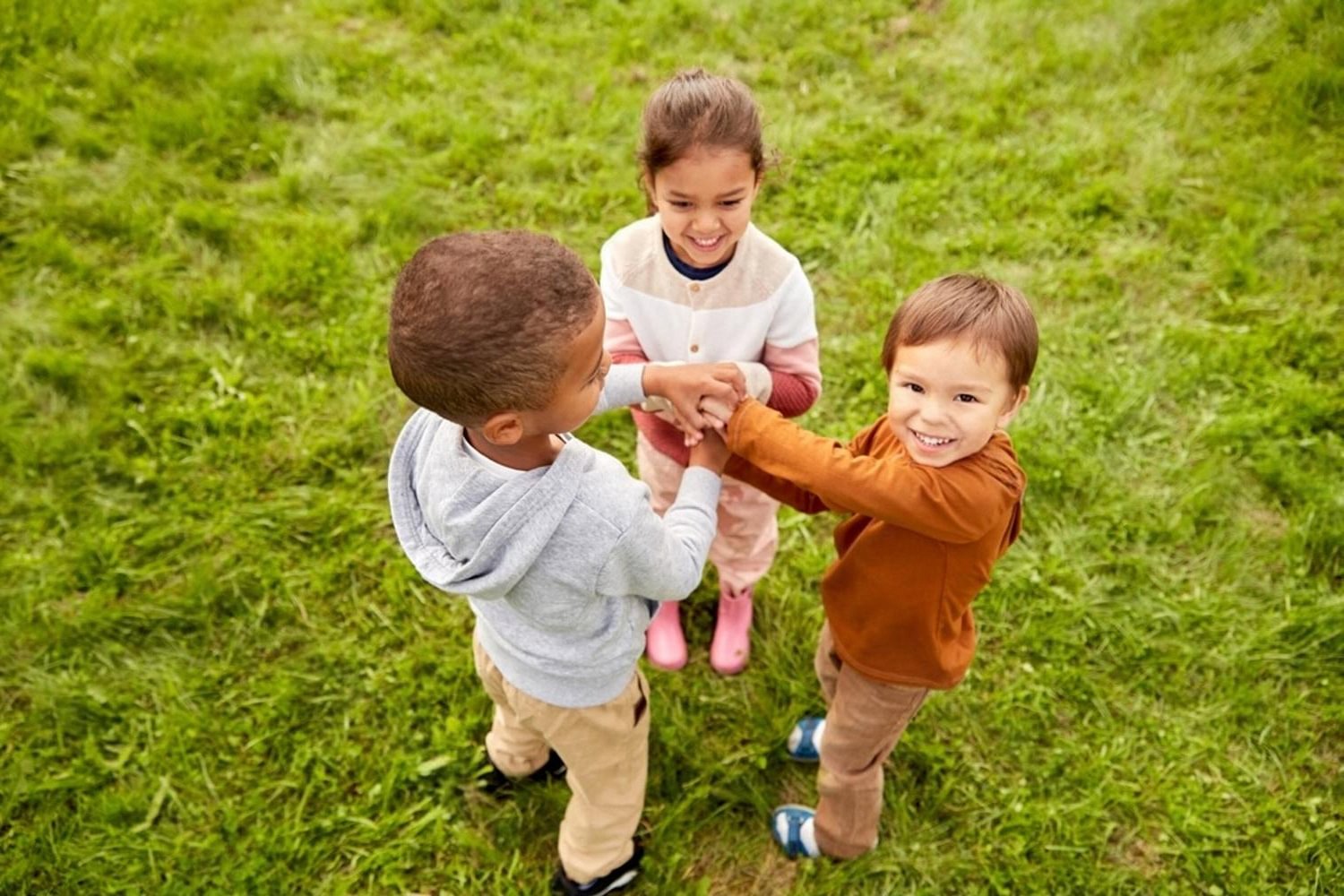 Three young children holding hands and playing in a circle on grass